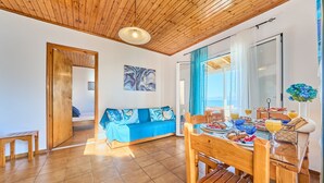 Ein Zimmer mit Holzbalkendecke, einem Sofa mit blauen Kissen, einem Esstisch mit Stühlen und Blick durch eine Glastür.