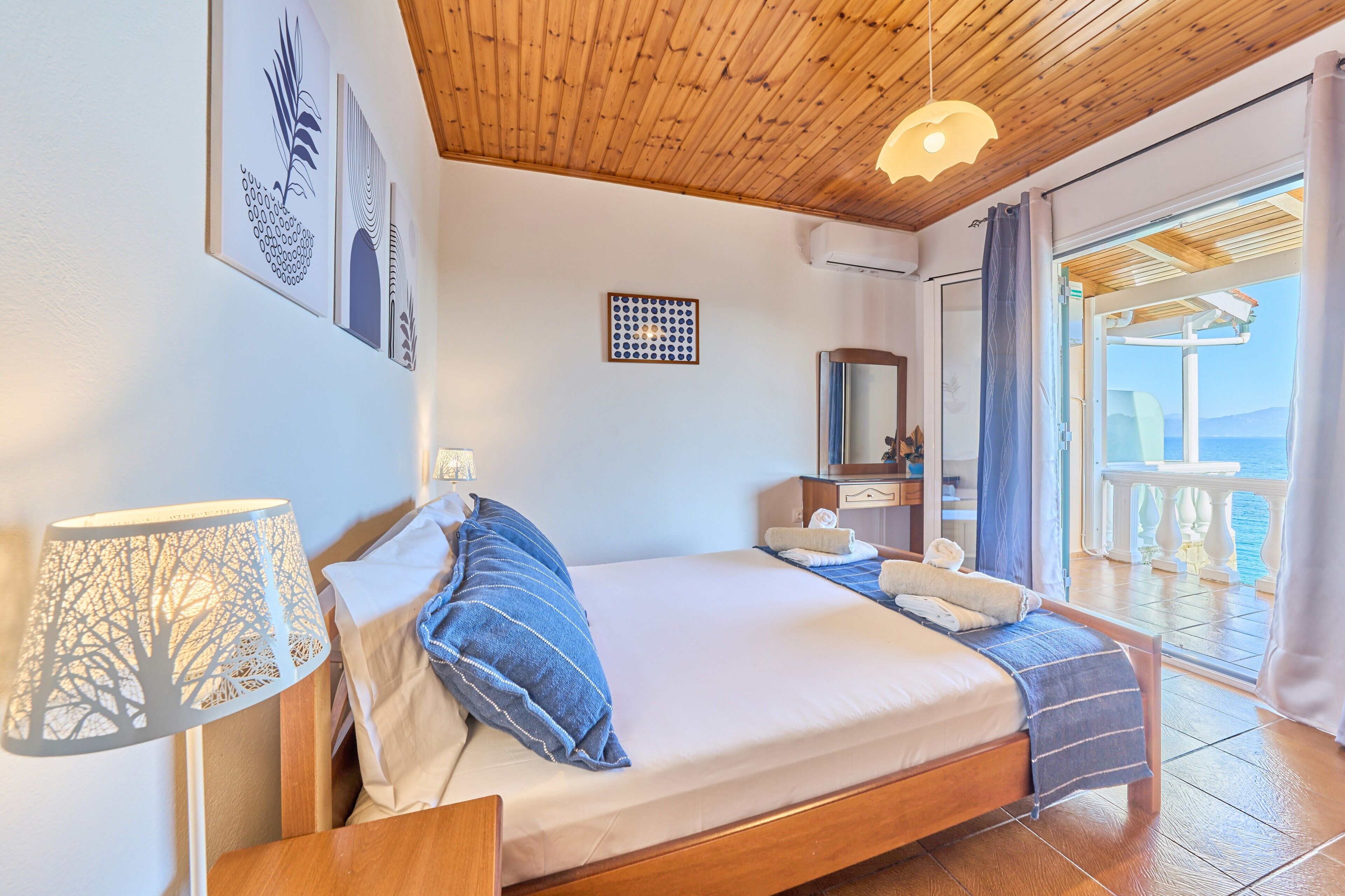 Ein Schlafzimmer mit Holzboden, einem Bett mit blauer Bettwäsche, einem Nachttisch, einer Lampe, einem Spiegel und Meerblick vom Balkon.