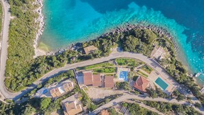 Luftaufnahme eines Anwesens an der Küste mit einem Pool, mehreren Gebäuden und einer Straße.