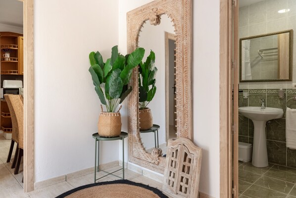 A hallway with a large ornate mirror, potted plants, a wicker chair, and a view into a bathroom with a sink and mirror. - Holiday Home 'Susana' with Private Pool, Private Terrace and Wi-Fi (Santa Eulària des Riu)