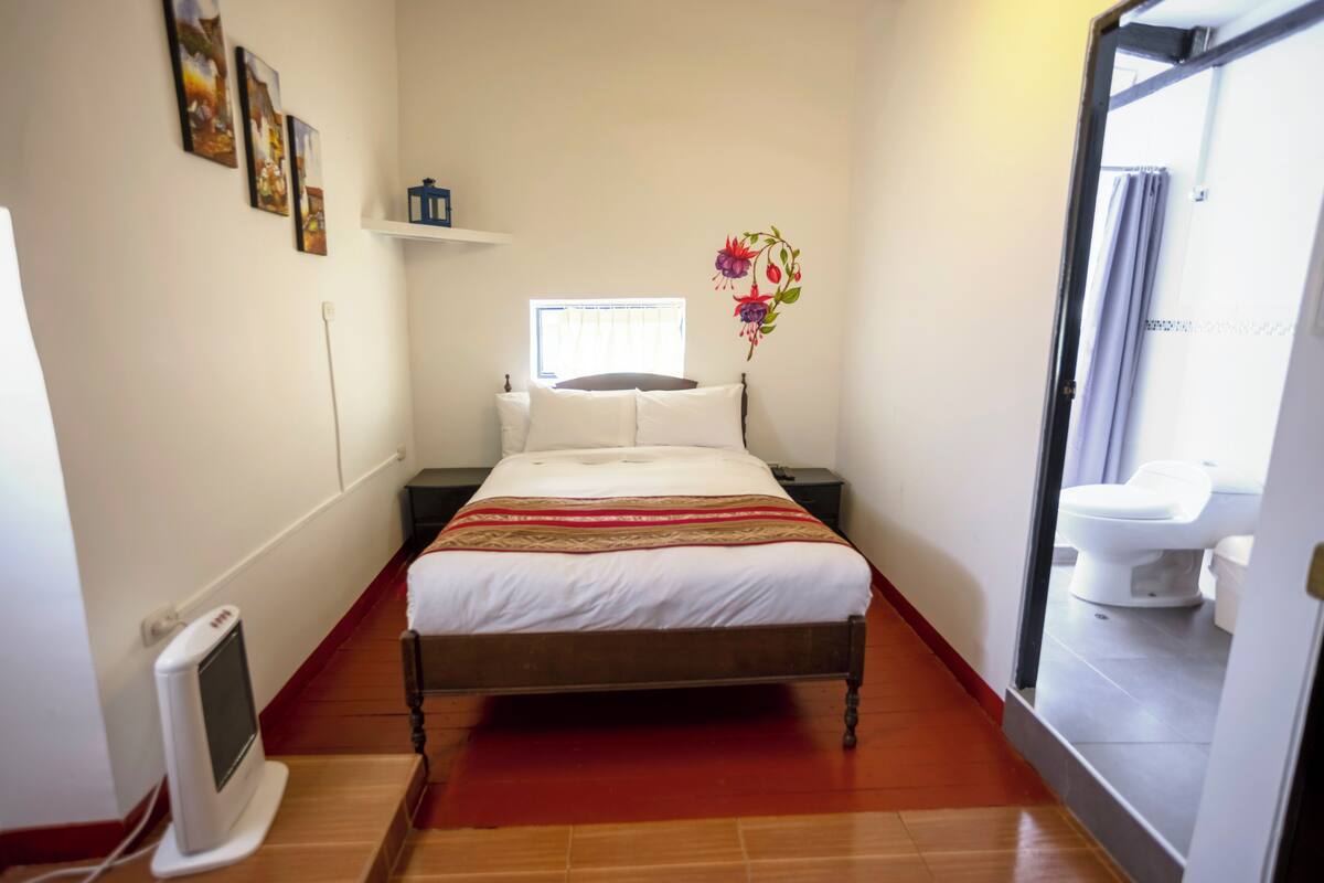 Une chambre à coucher avec un lit, des tables de chevet, un téléviseur fixé au mur, une fenêtre et une salle de bain visible à travers une porte ouverte.