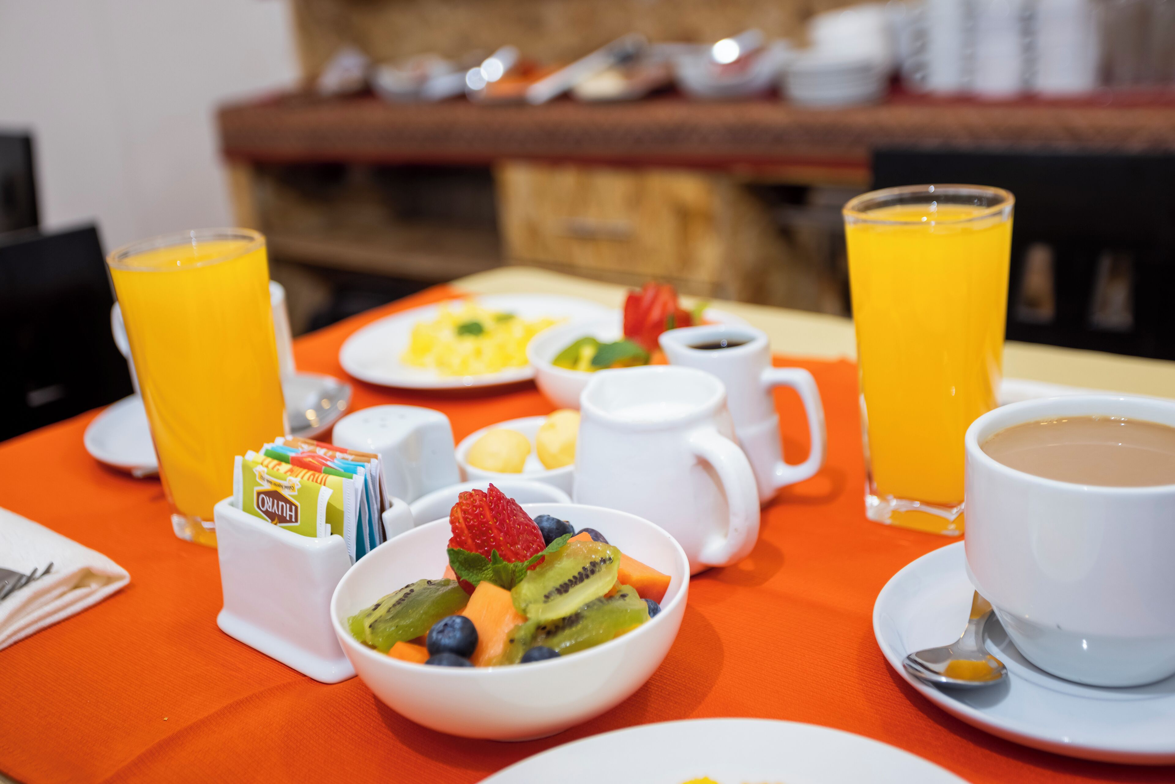 Une table de petit-déjeuner avec un saladier de fruits, deux verres de jus d’orange, une tasse de café et un petit pot de yaourt.