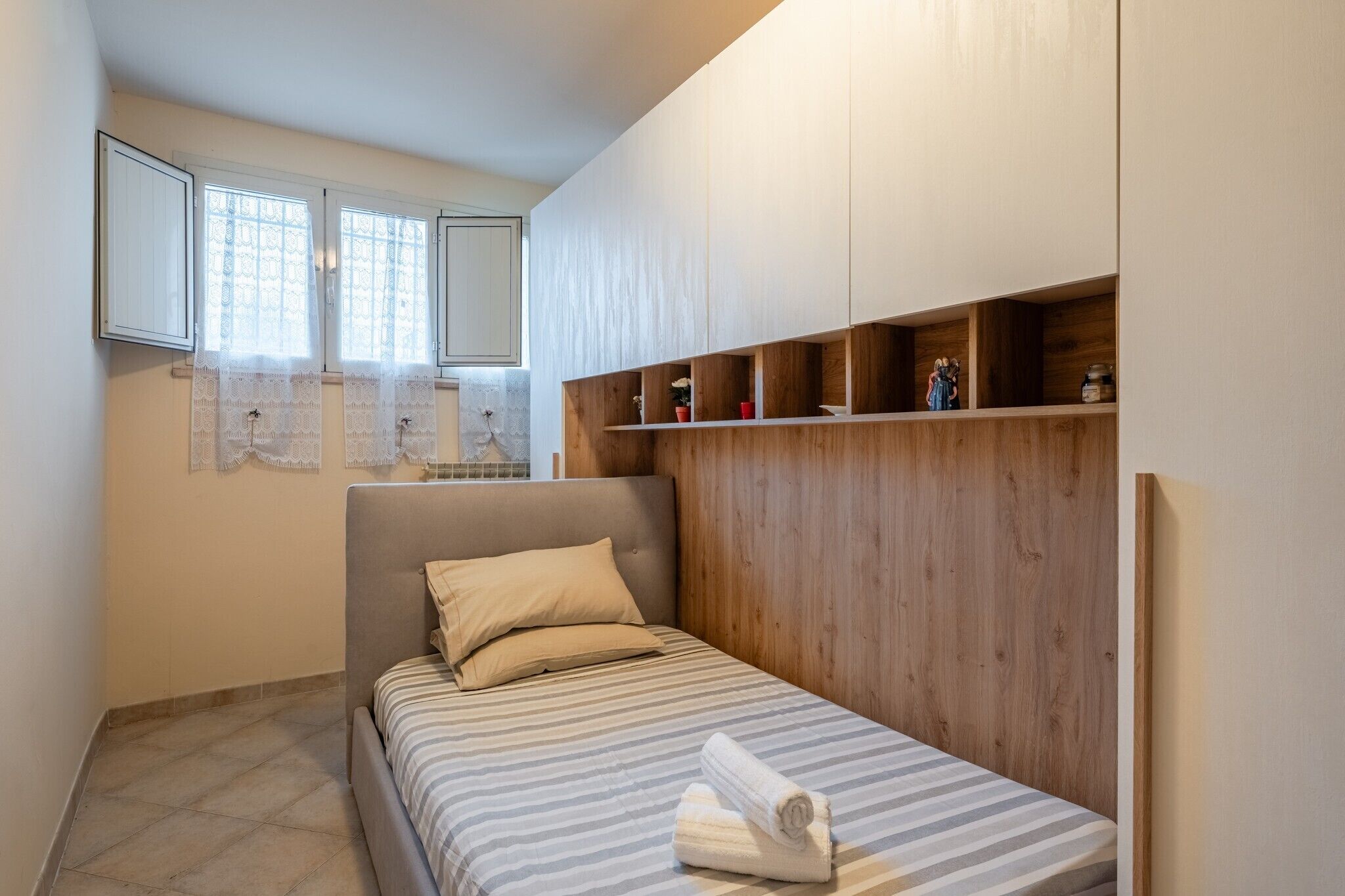 A bedroom with a bed, a headboard, a window with curtains, and a wooden shelf with items.