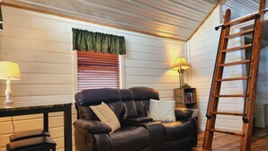 A cozy living room with a leather sofa, a wooden ladder, a table with a lamp, and a bookshelf. - Cabin of Western Skies:  loft, Wi-Fi , and tiny home living-close to OSU (Perkins)