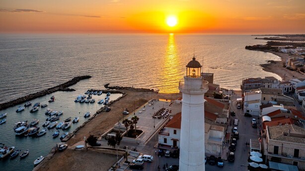 Un pueblo costero al atardecer con un faro destacado, un puerto deportivo con barcos y una combinación de edificios residenciales y comerciales.