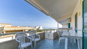 Un balcón con mesa y sillas, vista al mar y un edificio con un persiano verde.