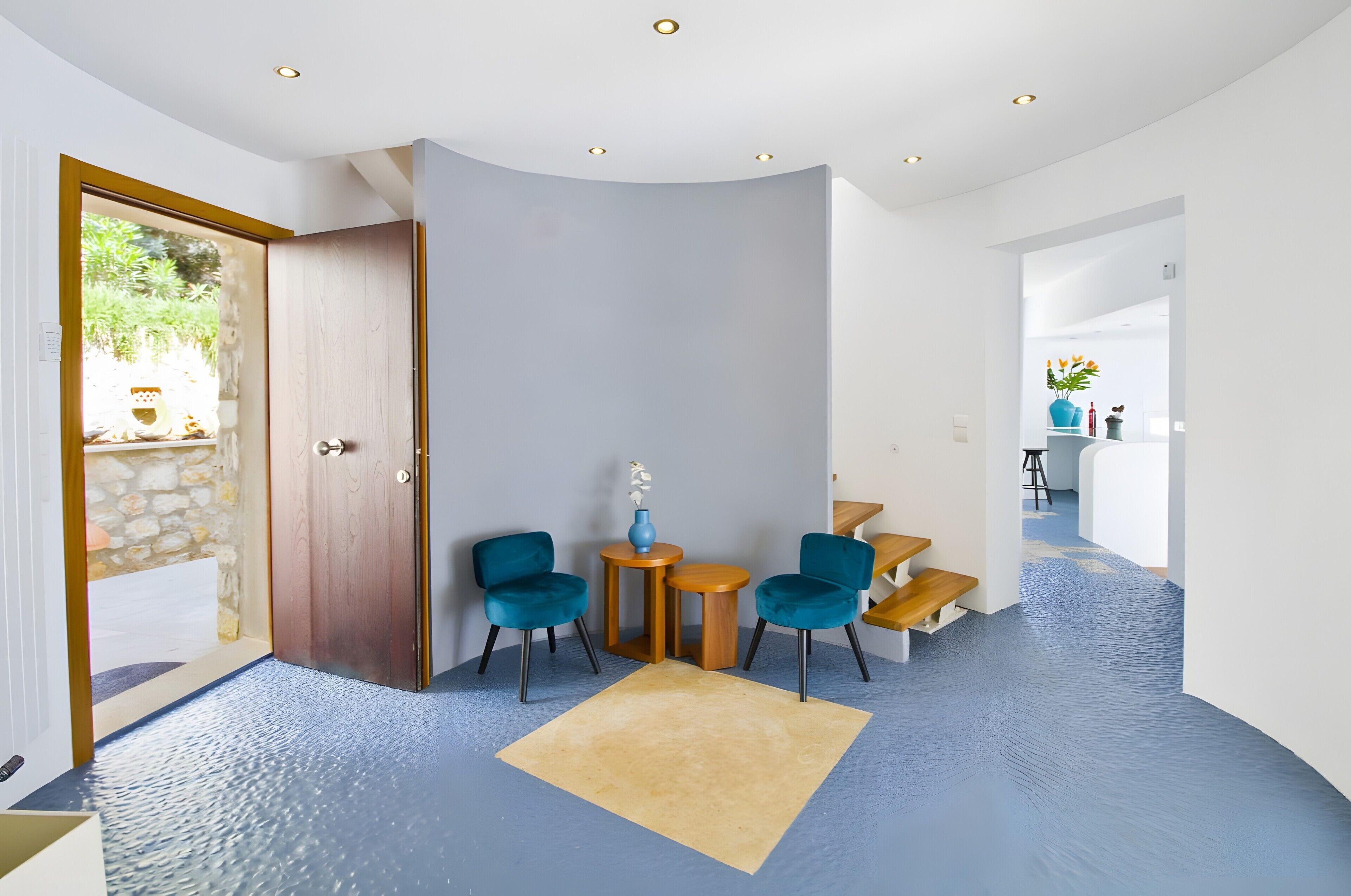 A modern interior with a curved ceiling, a wooden door, two blue chairs, a small round table, and a beige rug.
