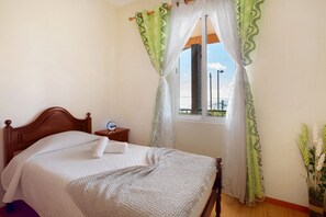 A bedroom with a wooden bed, a bedside table, a window with curtains, and a view of the sky.