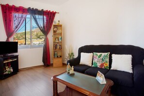 A living room with a sofa, a coffee table, a TV stand, a bookshelf, and a window with curtains.
