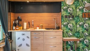 Une cuisine compacte dotée d’un plan de travail en bois, d’un petit réfrigérateur orné d’un autocollant représentant un chat, d’un évier et d’un mur décoré d’un motif de feuilles tropicales.