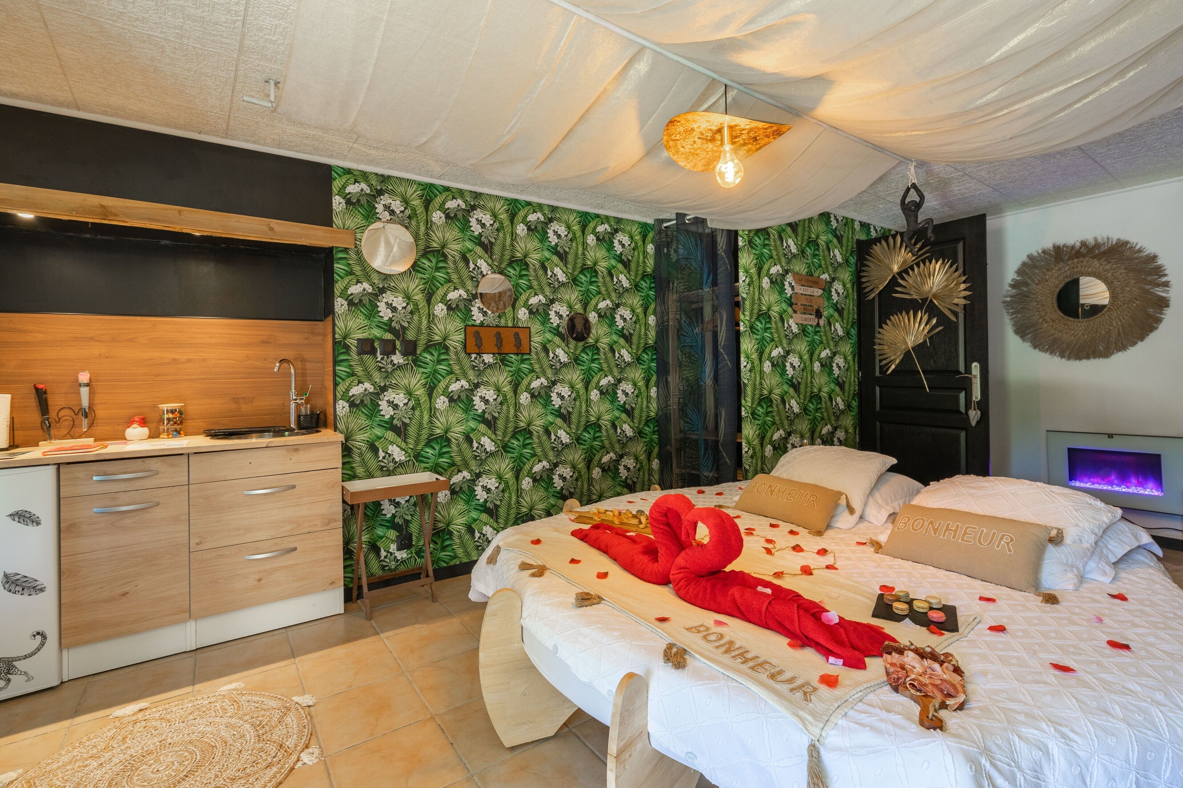A bedroom with a bed, a wooden dresser, a vanity, and a wall with tropical wallpaper.