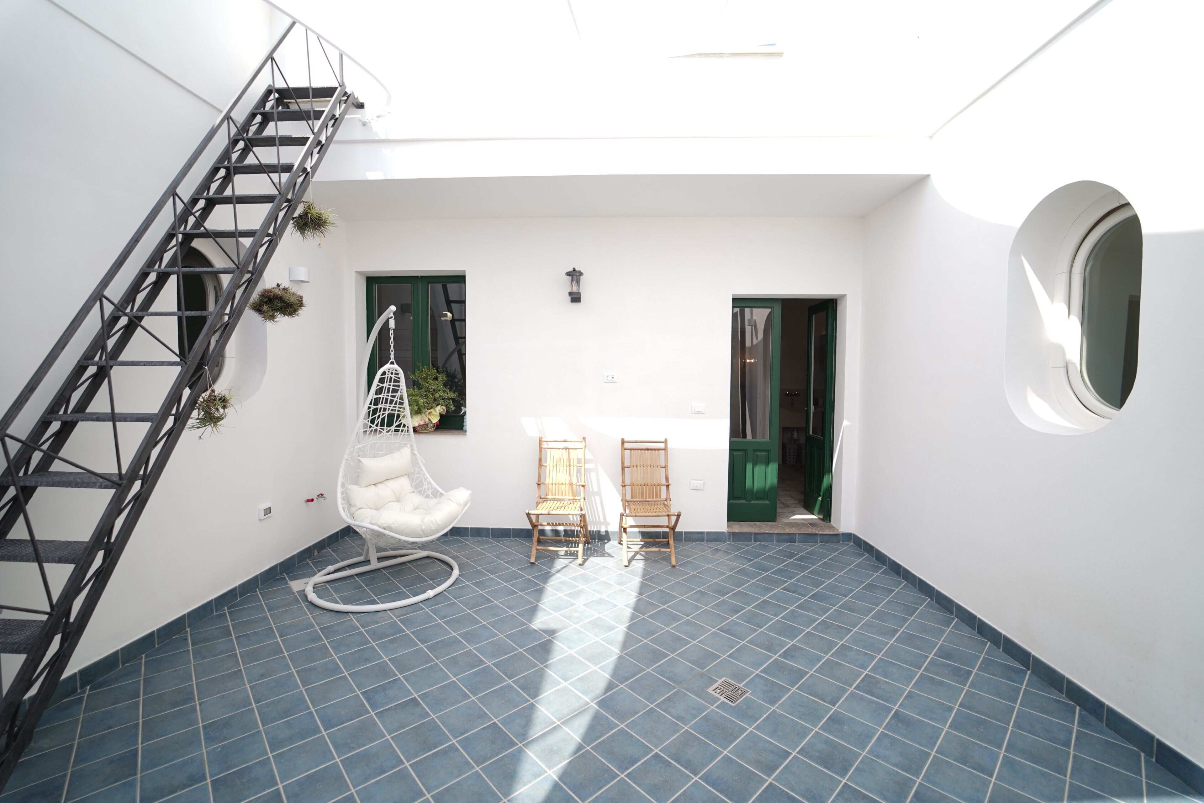 Une pièce avec un sol carrelé de carreaux bleus, une chaise blanche, deux chaises en bois, une chaise suspendue blanche, un escalier en métal et une fenêtre ronde.