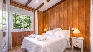 A bedroom with a wooden paneled wall, a large window, a bed with a quilted bedspread, two bedside lamps, and a nightstand with drawers. - Country House 'El Amanecer' with Mountain View, Wi-Fi and Air Conditioning (Motril)