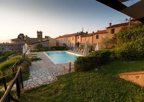A pool area with stone flooring, lounge chairs, and umbrellas, surrounded by greenery and traditional buildings. - Apartment 'Il Conte' with Shared Pool and Shared Garden (Massarosa)