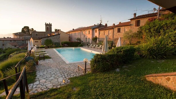 A pool area with stone flooring, lounge chairs, and umbrellas, surrounded by greenery and traditional buildings. - Apartment 'La Castellana' with Shared Pool, Wi-Fi and Air Conditioning (Massarosa)