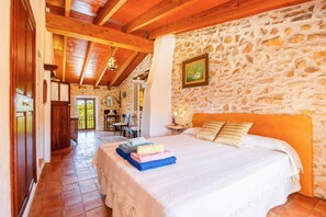 Quarto com parede de pedra, vigas de madeira, cama com almofadas e vista para o exterior.
