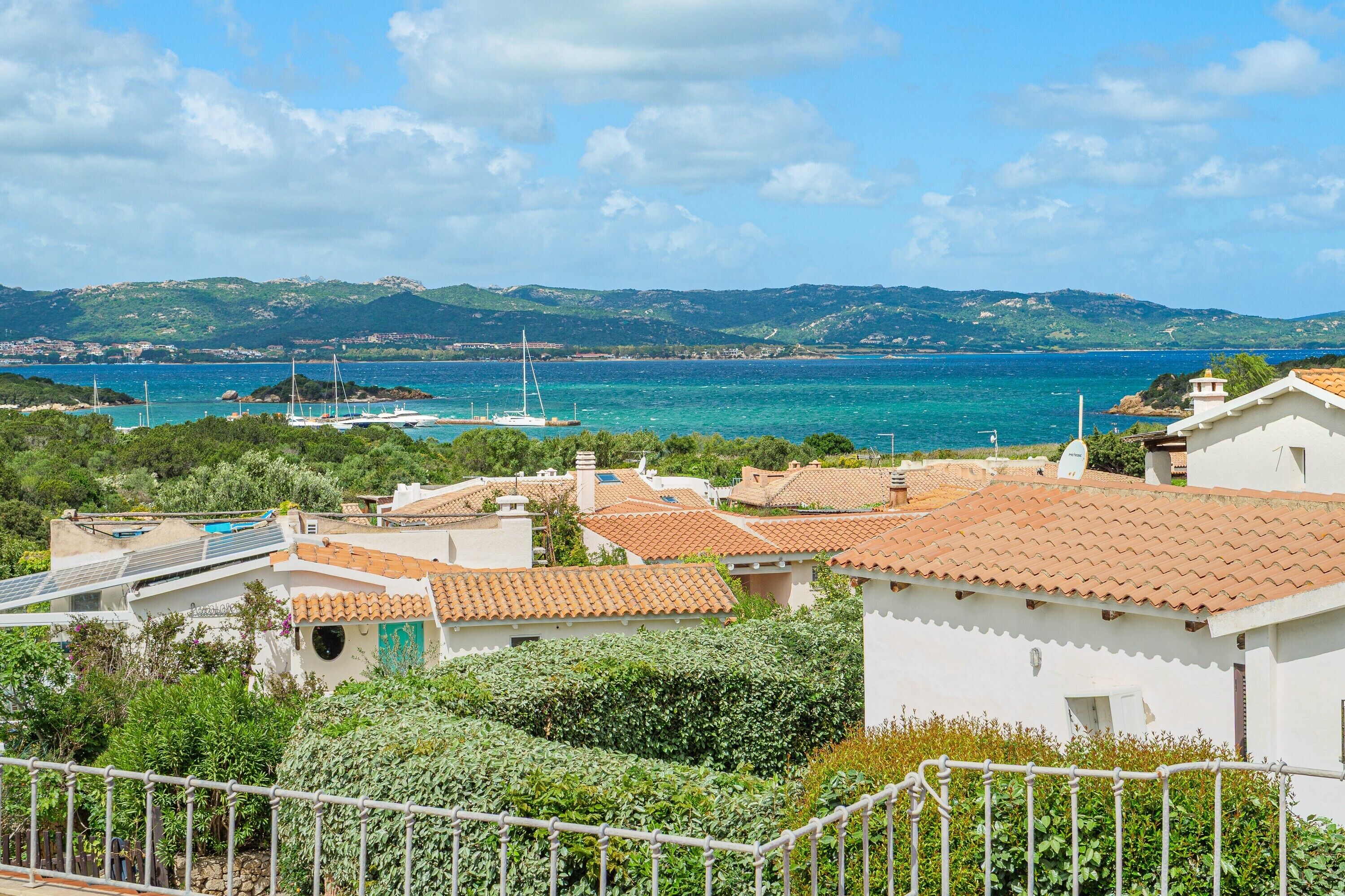 Uma vila costeira com edifícios de telhados em terracota, uma marina com veleiros e uma paisagem montanhosa ao fundo.