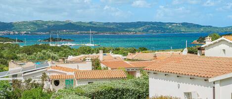 Uma vila costeira com edifícios de telhados em terracota, uma marina com veleiros e uma paisagem montanhosa ao fundo.