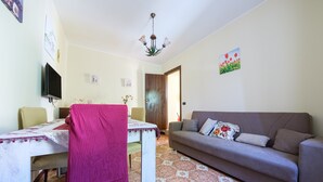 A living room with a sofa, a dining table with chairs, a TV mounted on the wall, and a patterned floor.