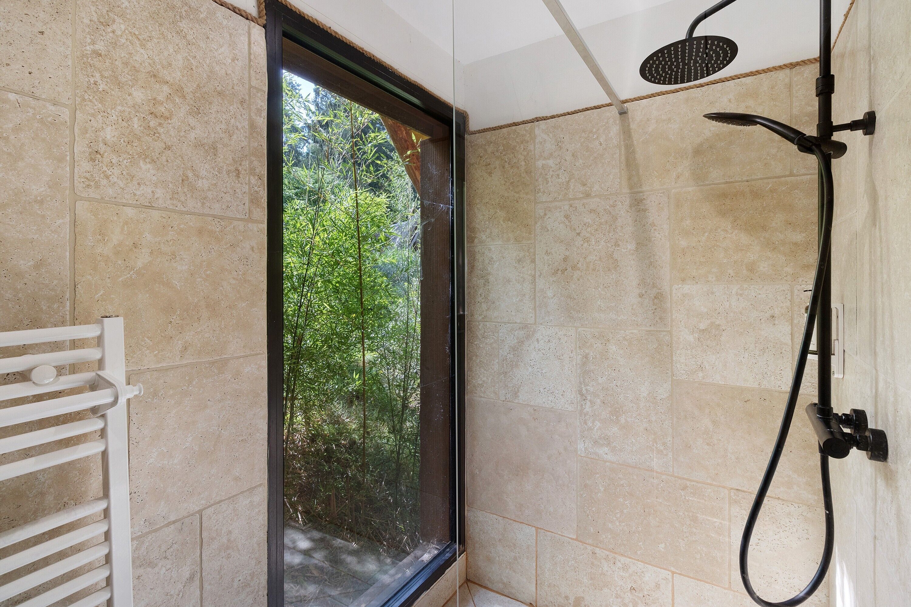 Ducha con cabezal de ducha de mano, ventana con vistas a zonas verdes y un perchero para toallas.