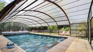 A covered swimming pool with a glass ceiling, surrounded by lounge chairs and a wooden deck.