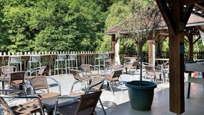 Een terras buiten met rieten stoelen en tafels, omgeven door een houten omheining en bomen.