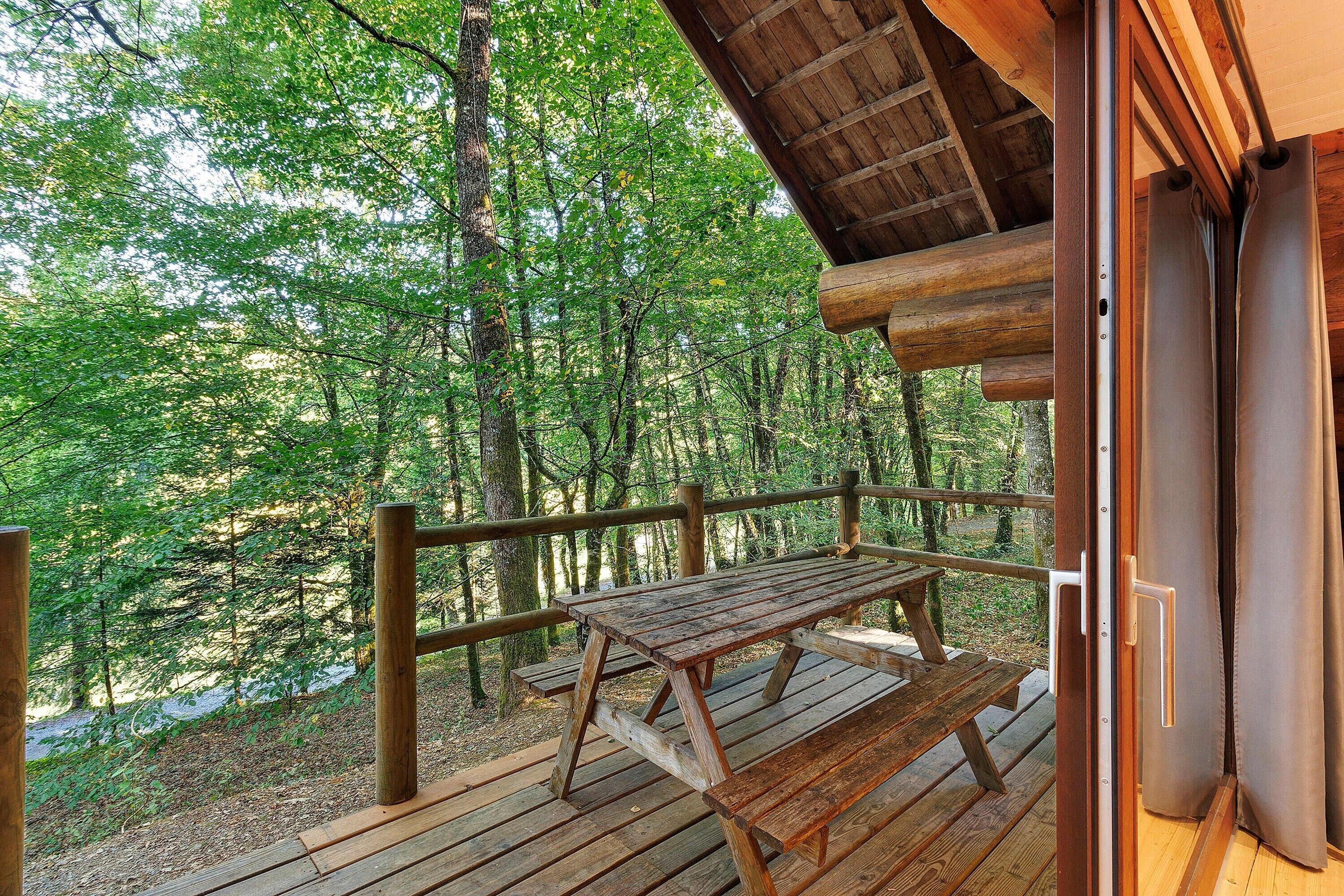 Une terrasse en bois avec une table de pique-nique, entourée d’une balustrade en bois, donnant sur une zone boisée.