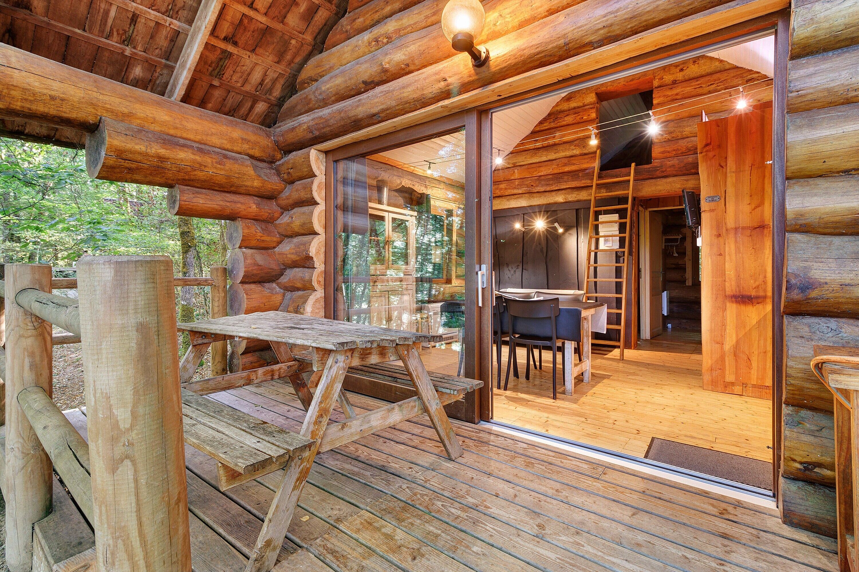 Une cabane en rondins dotée d’une terrasse en bois, d’un coin repas avec une table et des chaises, et d’une porte vitrée donnant sur l’intérieur.