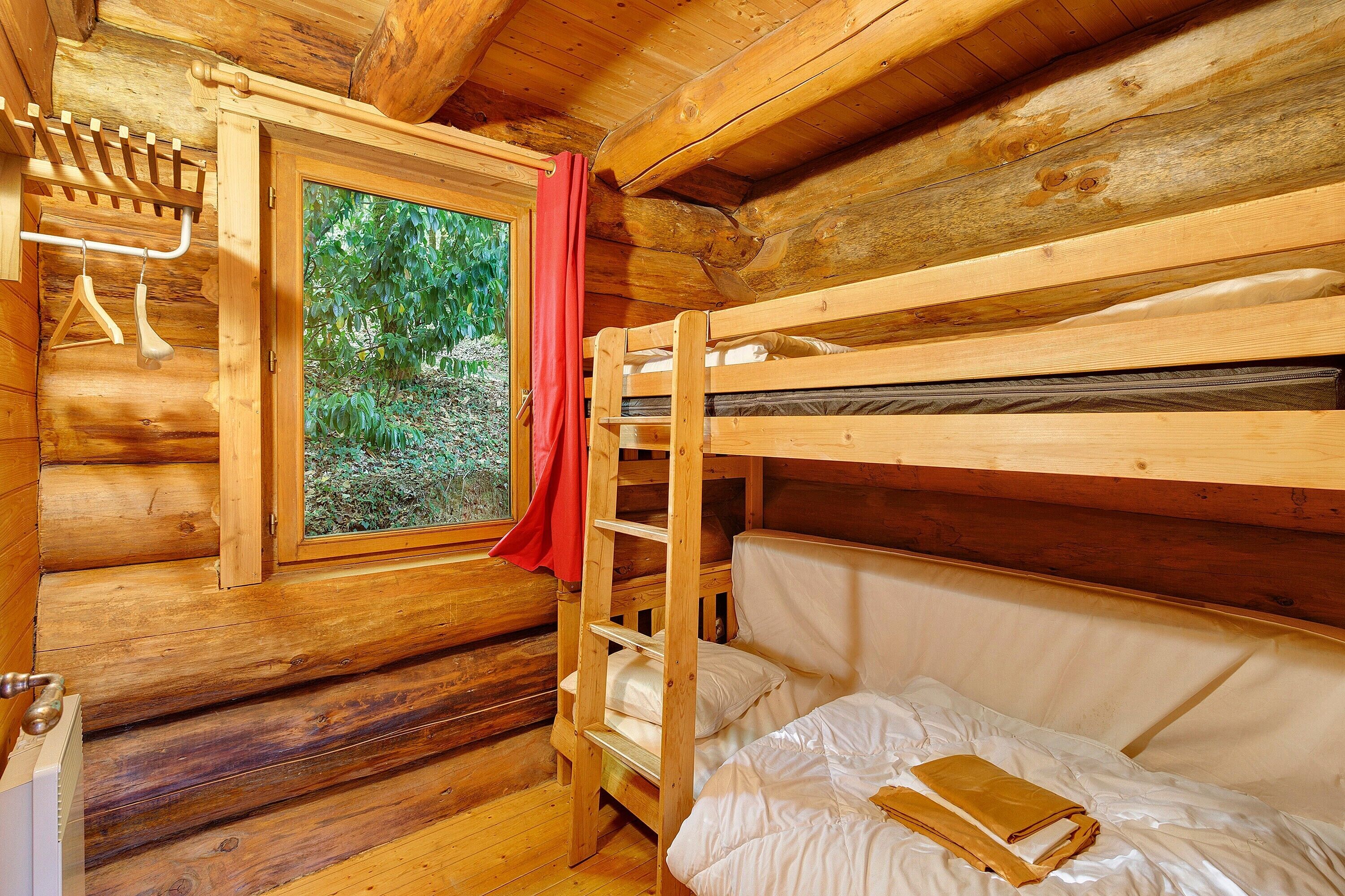 A wooden cabin room with a bunk bed, a window with a red curtain, and a wooden door with a towel rack.