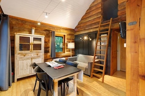 A cozy cabin interior with a dining table, chairs, a sofa, a ladder, and a cabinet with a mesh door.