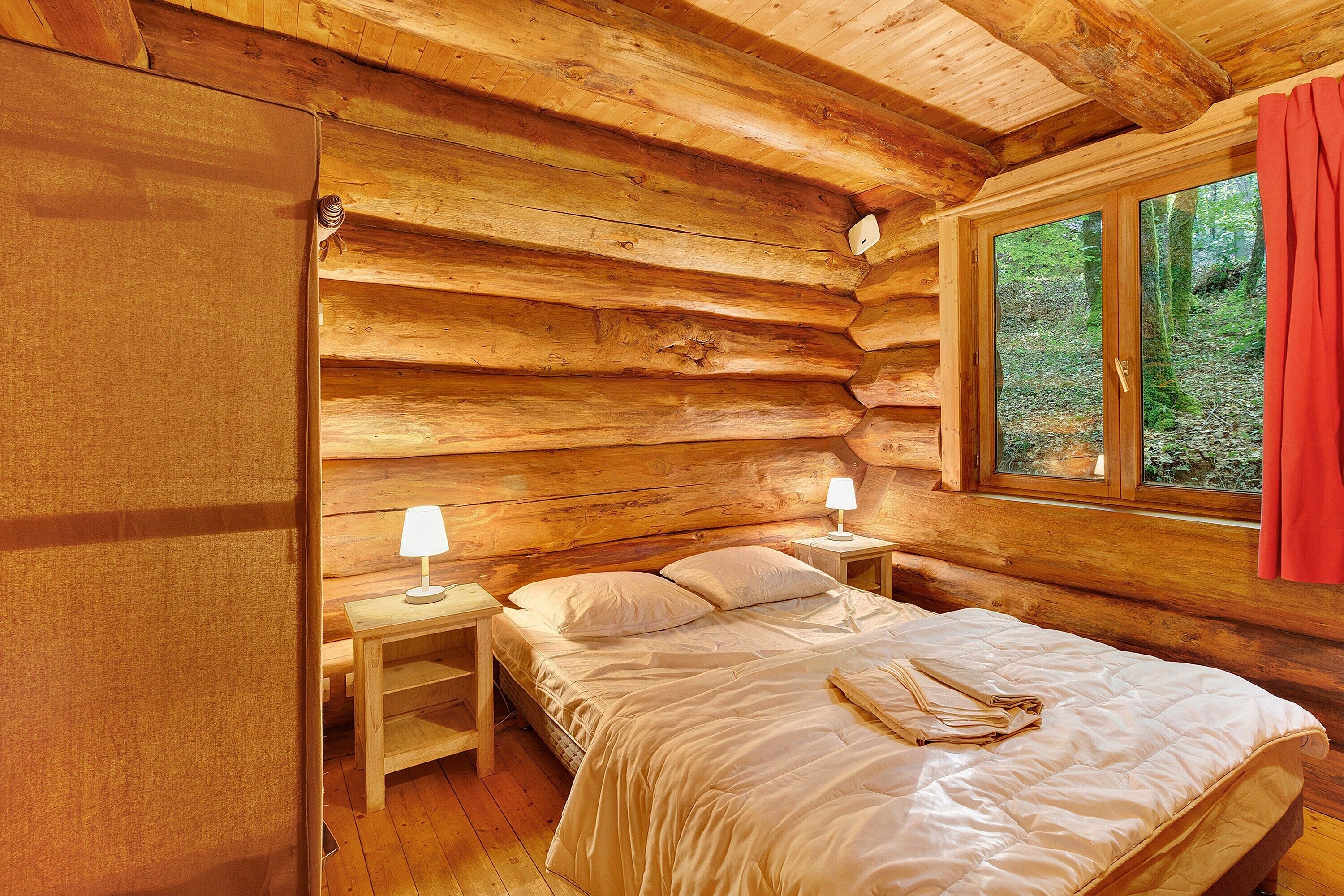 Une chambre à coucher située dans une cabane en rondins, avec un lit, deux tables de chevet et une fenêtre ornée d’un rideau rouge.