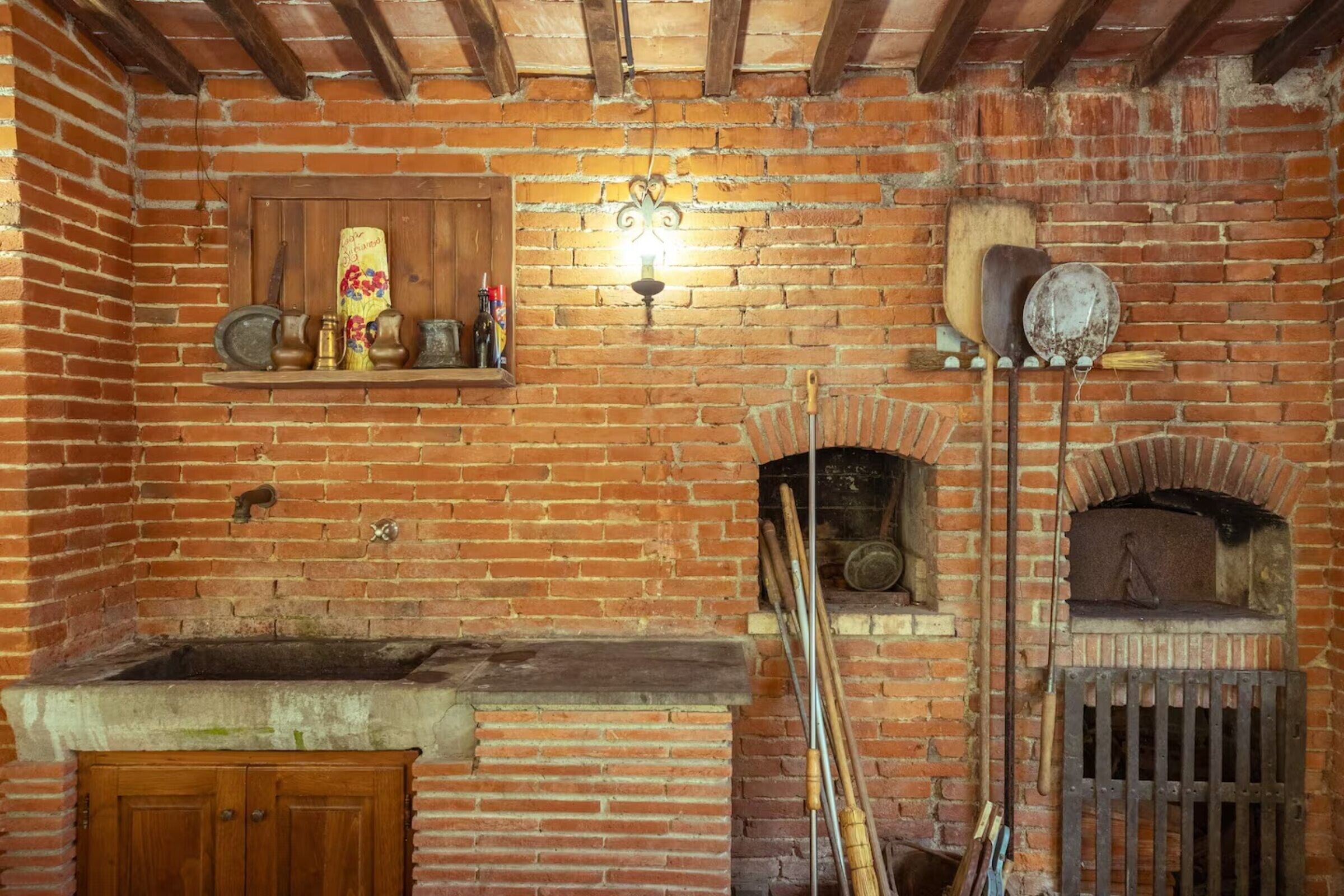 A rustic kitchen with a brick wall, wooden beams, a hanging light fixture, and a brick oven.