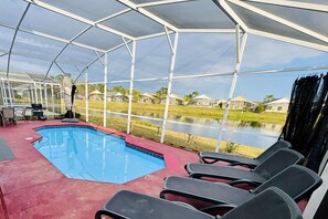Zona de piscina exterior cubierta con sillas de descanso, una pequeña mesa y vistas a un lago y casas al fondo.