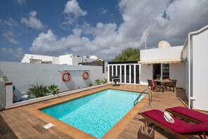Una piscina en la azotea con agua azul cristalina, rodeada de un suelo de madera, un área de descanso con cojines rojos y un edificio blanco al fondo.