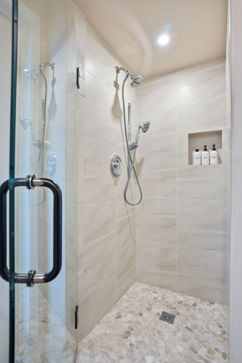 Un área de ducha con puerta de vidrio, un cabezal de ducha manual, un cabezal de ducha empotrado en la pared y un estante pequeño con artículos de aseo.