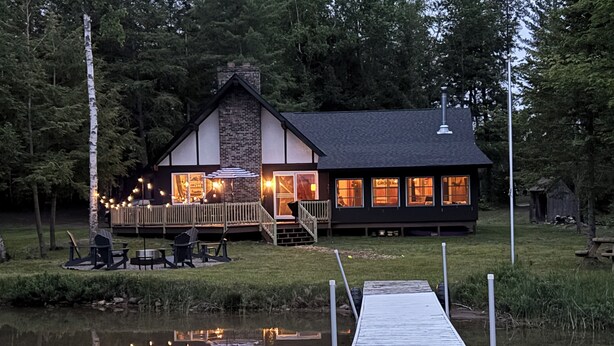 A lakeside cabin with a dock, deck seating, and a visible interior with warm lighting.