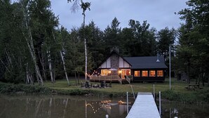 A lakeside lodge with a dock extending into the water, featuring a deck with outdoor seating and a lit interior.