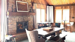 A cozy room with a stone fireplace, wooden ceiling beams, a dining table with chairs, and a window with curtains.