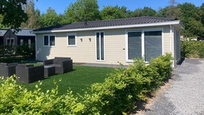 Ein eingeschossiges Haus mit flachem Dach, einer Veranda mit Sitzbank und einem kleinen Vorgarten mit Rasen und Büschen.