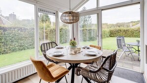 Un coin repas avec une table ronde dressée pour quatre personnes, entourée de chaises en osier, d’un luminaire suspendu et offrant une vue sur un jardin luxuriant grâce à de grandes fenêtres.