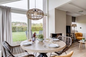 A dining area with a round table set for four, surrounded by wicker chairs, a hanging light fixture, and a view of the outdoors through large windows.