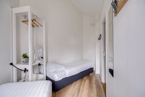 Una habitación pequeña y limpia, con una cama individual, un armario blanco con espacio para colgar ropa, un estante con una planta y un espejo en la pared.