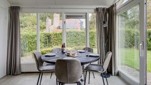 A dining area with a round table set for six, surrounded by chairs, large windows with curtains, and a view of a garden and buildings.