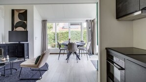 A modern hotel room with a dining area, a flat-screen TV, a kitchen, and a large window offering a view of greenery.