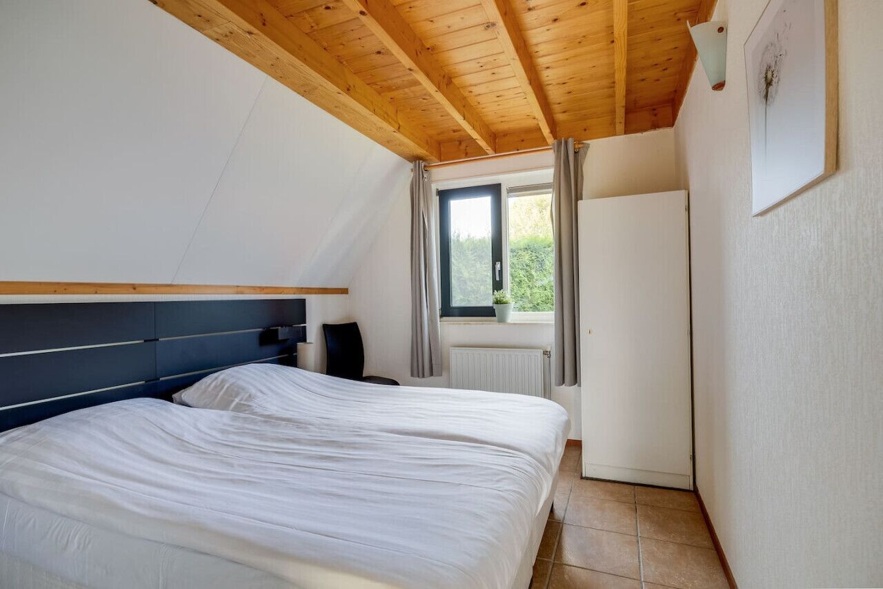Camera da letto con soffitto in legno, un letto, un comodino, una finestra con tende e un termosifone.