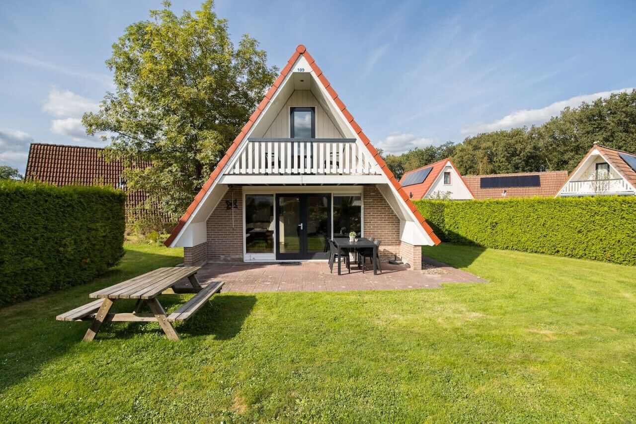A triangular-roofed building with a balcony, a patio with a table and chairs, and a well-maintained lawn.