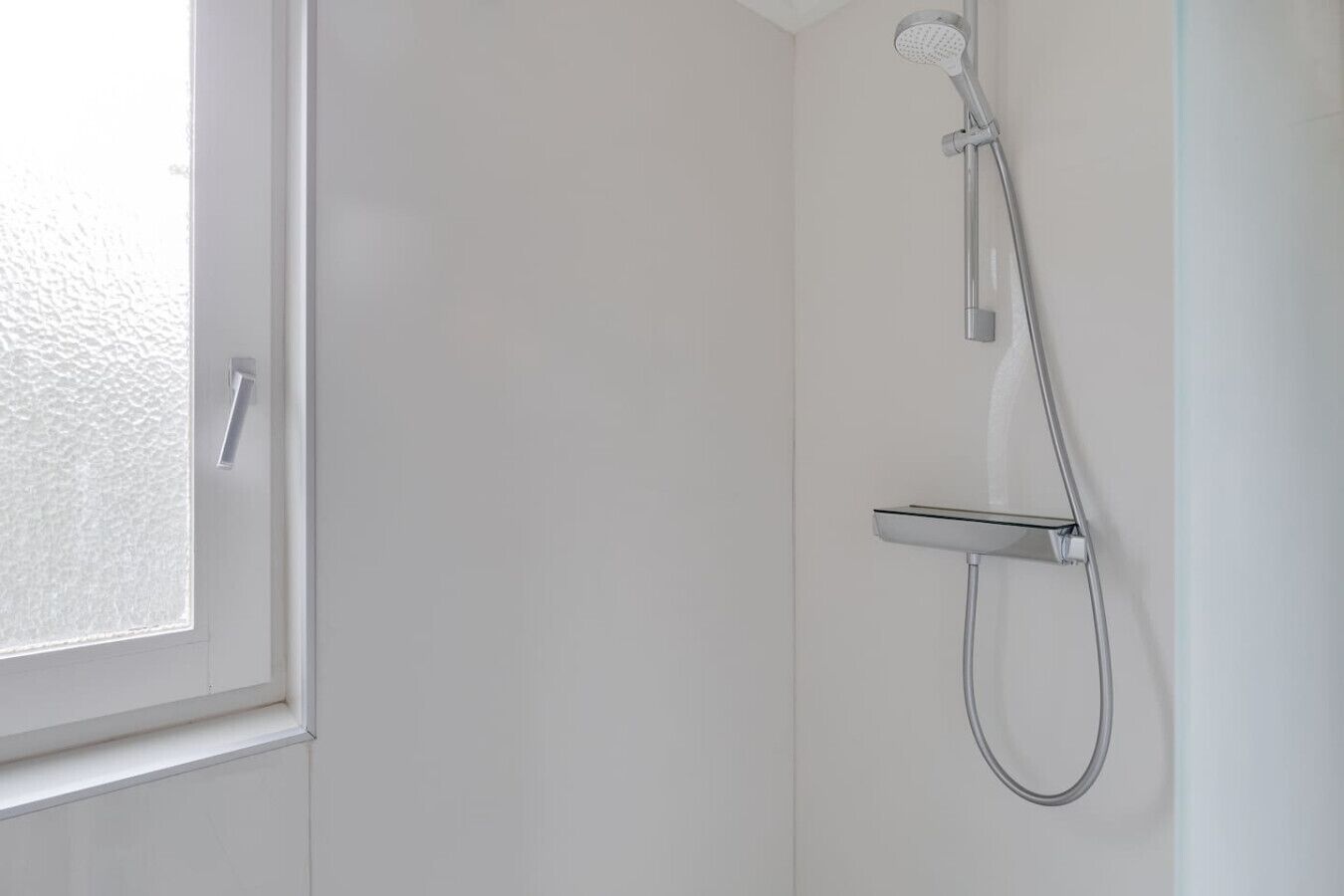 Ducha con cabezal de ducha de mano y panel de control en la pared, en un cuarto con paredes blancas y una ventana.