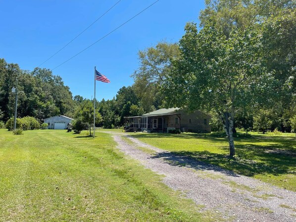 A rural setting with a gravel path leading to a brick house with a porch. An American flag is visible in the background. - Beautiful brick home w/ large porch and 3 bedrooms (Bowman)