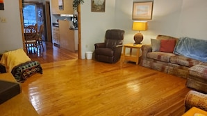 A living room with a wooden floor, a brown leather armchair, a patterned sofa, and a small table with a lamp. - Beautiful brick home w/ large porch and 3 bedrooms (Bowman)