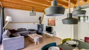 A modern living room with a sofa, coffee table, and dining table. There is a TV, a fireplace, and a hanging light fixture.